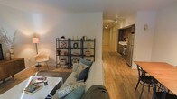 This single-floor residence offers a thoughtfully designed layout that blends comfort and functionality. The living area is warm and inviting, anchored by a neutral-toned sofa adorned with accent pillows, a low-profile coffee table, and an open metal shelving unit that provides both storage and display space for decorative accents. Recessed lighting illuminates the space, complementing the rich hardwood flooring that runs throughout the unit and ties each area together seamlessly.

A wide corridor connects the living area to the kitchen, which is equipped with stainless steel appliances including a refrigerator and range. Dark cabinetry contrasts with lighter countertops and walls, giving the kitchen a sleek, modern aesthetic. The open sightlines from the living space into the kitchen enhance the sense of openness and make the floor plan feel expansive.

The consistent warm-toned hardwood flooring, clean white walls, and recessed and track lighting throughout contribute to a bright, cohesive ambiance. The overall design balances practicality with a contemporary residential aesthetic, making it a comfortable and visually appealing living environment.