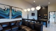 This single-floor space is designed with entertainment and socializing in mind, offering a cohesive blend of modern aesthetics and functional design. The lounge area anchors the space with a substantial dark leather sectional sofa adorned with decorative pillows, providing ample seating for guests. A striking large-scale abstract artwork in oceanic blues and greens commands attention on the wall, adding a bold visual focal point.

Overhead, a row of four sculptural pendant lights with organic, feathery white shades cascades from the ceiling, serving as both a functional lighting source and an artistic statement. The elevated ceiling height enhances the sense of openness throughout the space.

A sleek dark bar counter with barstool seating runs along the mid-section of the room, creating a natural division between the lounge and the entertainment zone. A wall-mounted flat-screen television is positioned conveniently nearby. Beyond the counter, a classic arcade machine and what appears to be a billiards table extend the space's entertainment offerings. The overall ambiance is sophisticated yet inviting, with a dark, moody color palette balanced by strategic accent lighting.