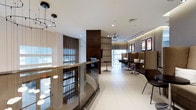 This sophisticated single-floor lounge space exudes a polished, contemporary aesthetic through its carefully curated design elements. The interior is anchored by large floor-to-ceiling windows dressed with floor-length brown drapes, allowing natural light to fill the space while maintaining an air of refined privacy. Crisp white tiled flooring runs throughout, creating a bright and clean foundation that contrasts elegantly with the warm, dark-toned furniture and architectural accents.

A distinctive curved glass and brass railing feature adds a striking architectural focal point, lending a sense of openness and visual flow across the space. The lounge area is well-appointed with high-back booth-style seating upholstered in rich brown tones, complemented by bistro-style tables and chairs that create intimate gathering zones. A prominent dark wood structural column and shelving unit serves as a stylish room divider, contributing to the space's layered layout.

Lighting is thoughtfully layered, combining recessed ceiling fixtures, elegant pendant clusters, and a ceiling fan, all working together to create a warm and inviting ambiance. Framed wall art adds a curated touch to the seating area, while the overall neutral palette of cream, taupe, and chocolate brown creates a cohesive, upscale environment well-suited for relaxed social or professional gatherings.