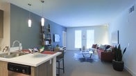 This single-floor residence showcases a thoughtfully designed open-concept layout that integrates the kitchen and living areas into one cohesive space. The kitchen features a sleek white breakfast bar with seating, complemented by pendant lighting that adds a contemporary touch. Rich wood-toned cabinetry and a lean ladder shelf unit provide functional storage while enhancing the room's modern aesthetic.

The accent wall, painted in a cool slate blue, creates a striking contrast against the crisp white ceiling and surrounding walls, adding depth and visual interest to the space. A wall-mounted flat-screen television is positioned on the feature wall, flanked by the decorative shelving unit displaying books and décor items.

The living area is carpeted in neutral tones, grounding the space and providing comfort underfoot. A cowhide-style area rug defines the seating zone, where a burgundy sofa and light gray accent chair are arranged around a contemporary glass and metal coffee table. Large windows dressed with white blinds allow generous natural light to fill the room, creating a bright and welcoming ambiance throughout.