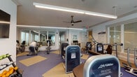 Located at 3650 RCA Blvd in Palm Beach Gardens, Florida, this fitness center offers a comprehensive, well-equipped environment designed to support a full range of workout routines. The main exercise floor features an extensive selection of Life Fitness strength machines—including leg extension, seated leg curl, lat pulldown, and cable systems—alongside cardio equipment such as treadmills, upright and recumbent bikes, and ellipticals. The space is finished with a durable two-tone blue and tan rubberized floor and illuminated by long linear LED fixtures overhead, complemented by ceiling fans that promote air circulation throughout. Large windows along one wall allow natural light to filter in and provide views of the surrounding greenery, enhancing the open and airy atmosphere. A wall-mounted television and a dumbbell rack complete the main workout area. Adjacent to the gym floor is a dedicated movement and wellness studio featuring warm wood-look flooring, teal-blue accent walls, floor-to-ceiling windows with lush outdoor views, mirrored walls, and rows of upholstered seating chairs—ideal for group classes or instructional sessions. The facility also includes a clean, tiled locker room area with a shower stall and a wooden bench, providing convenient amenities for post-workout use. Fitness center guidelines are posted on-site for member reference.