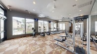 Located at 14700 Woodson Park Drive in Houston, Texas, this well-equipped fitness center offers a comprehensive workout environment across a single, open floor. The space is lined with large windows on multiple walls, flooding the interior with abundant natural light while providing views of the surrounding community. Ceiling fans and recessed lighting complement the bright, airy atmosphere.

The patterned carpet tiles in earthy tones of tan and gold add a distinctive visual texture to the floor throughout the space. A full wall of mirrors on one side visually expands the room and is essential for proper form during strength training exercises.

The cardio zone features a row of treadmills and elliptical trainers, as well as a stationary bike, all positioned near the windows to take advantage of natural light. Multiple wall-mounted flat-screen televisions are installed throughout for entertainment during workouts. The strength training area is equipped with a multi-station cable machine, a weight bench, and a well-stocked dumbbell rack ranging across various weights. A functional training corner includes medicine balls, stability balls, and a mat area for stretching and bodyweight exercises.

Entry to the fitness center is through a set of double glass doors. Framed artwork on the walls adds a polished, community-amenity feel to this thoughtfully designed workout facility.