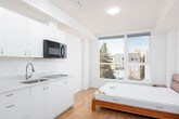 This well-designed studio unit offers a thoughtfully arranged open-plan layout that seamlessly combines living, sleeping, and cooking functions within a single, efficiently organized space. Light hardwood flooring runs throughout, complementing the clean white walls and ceiling to create a bright, airy atmosphere.

The sleeping area anchors one end of the unit and is positioned to take full advantage of large, floor-to-ceiling windows that frame views of the surrounding neighborhood and bring in generous amounts of natural light. A wall-mounted microwave and a compact kitchenette with a cooktop and under-counter cabinetry sit adjacent to the sleeping area, providing convenient access to essential cooking facilities.

Toward the entry, a dedicated kitchen zone features ample white cabinetry — including tall storage columns and multi-drawer lower units — alongside a full-size refrigerator, a sink, and additional counter space, offering impressive storage capacity for a studio of this scale.

A cozy dining nook accommodates a warm-toned wooden table and chairs, providing a defined space for meals or a work-from-home setup. The entry corridor is framed by the light wood front door with a modern lever handle. The overall design is clean and contemporary, with recessed and flush-mount lighting throughout, making this a functional and comfortable living space.