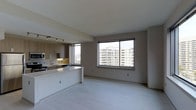 This contemporary single-floor space is designed with an open-concept layout that seamlessly connects the kitchen and living areas, creating a bright and functional environment. The kitchen is outfitted with warm-toned flat-panel cabinetry, a light stone countertop with a waterfall peninsula, under-cabinet lighting, a stainless steel sink, and modern appliances including a built-in microwave. Track lighting overhead adds a practical yet stylish touch to the culinary space. A large picture window in the living area floods the space with natural light while framing sweeping views of the surrounding urban skyline and neighboring mid-rise buildings — a standout feature of this unit. The living area is generously proportioned, offering ample room for a variety of furniture arrangements. Light-toned wide-plank flooring runs throughout, contributing to the clean, modern aesthetic. The neutral warm-gray walls complement the overall palette, creating a cohesive and inviting ambiance. With its elevated vantage point, open design, and quality finishes, this space is well-suited for those seeking a modern urban lifestyle.