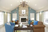 This single-floor community space is thoughtfully designed to feel both functional and inviting. The centerpiece of the lounge area is a striking floor-to-ceiling stacked stone fireplace with a classic white mantle, anchoring a vaulted ceiling that lends a sense of grandeur and openness to the room. The lounge is furnished with a mix of teal upholstered chairs and rich brown leather seating, all arranged atop a decorative area rug with bold botanical motifs. A chandelier adds an elegant finishing touch overhead, while teal accent walls and coordinating drapes tie the color scheme together cohesively. Large windows flood the space with natural light and offer views of the surrounding exterior.

Adjacent to the lounge, a dining or meeting area features a round dark wood table surrounded by striped upholstered chairs, alongside a work desk and seating nook, making the space suitable for both casual gatherings and professional use. The entry foyer is bright and accessible, with glass-paneled French doors, tile flooring, and a clean, welcoming aesthetic. A beverage station and a decorative convex mirror add practical and stylish touches to the workspace area. Light-toned tile flooring runs throughout, complementing the warm, neutral wall tones and creating a consistent, polished look across the entire space.