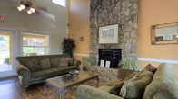 This single-floor space makes a strong architectural statement through its open, flowing layout and carefully chosen design elements. The centerpiece of the living area is a striking floor-to-ceiling natural stone fireplace, its rugged texture and earthy tones commanding immediate attention. Soaring ceilings amplify the sense of grandeur, with high-set windows drawing in natural light and further emphasizing the vertical scale of the room. Warm terracotta tile flooring runs throughout, lending a cohesive and inviting character to the space. The living area is generously proportioned, comfortably accommodating multiple seating arrangements centered around an ornate glass-top coffee table. Earthy, peach-toned walls complement the stone and tile palette, creating a warm and welcoming ambiance. A ceiling fan with integrated lighting provides both comfort and illumination. A glass-paneled entry door at the front floods the space with additional natural light and offers views of the outdoors. The layout transitions smoothly into a dining area and kitchen, visible toward the rear of the space, where cabinetry and appliances can be seen. The overall design balances rustic charm with comfortable, functional living, making this a distinctive and characterful space.