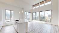 This contemporary single-floor residence showcases a thoughtfully designed open-concept layout bathed in natural light. The living and dining area is defined by dramatic double-height ceilings and an expansive wall of floor-to-ceiling windows and sliding glass doors, framing sweeping city skyline views and flooding the interior with daylight. Light-toned hardwood flooring runs throughout the space, complementing the clean, bright aesthetic.

The modern kitchen is a standout feature, centered around a large white waterfall island with an integrated sink. Sleek, high-gloss white cabinetry extends to the soaring ceiling, paired with a subtle tile backsplash and stainless steel appliances, including a refrigerator, range, and microwave. Elegant pendant lighting hangs above the island, adding warmth and visual interest.

The bedrooms offer generous proportions, elevated ceilings, and bright windows with city views. One bedroom includes a walk-in closet with built-in shelving. The bathrooms are finished in a clean, contemporary style — one features a double-sink vanity with dark wood cabinetry, a large mirror, and a soaking tub with white subway tile surround; a second bathroom offers a tub and wall-mounted fixtures. The overall ambiance is airy, bright, and refined, with a cohesive modern palette throughout.