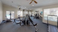 This single-floor fitness center is designed to provide a comprehensive workout environment for its users. The space is anchored by a robust multi-station cable and weight machine that serves as the centerpiece, complemented by a weight bench and a stationary exercise bike, offering a well-rounded range of strength and cardio training options. Full-length mirrors line one wall, visually expanding the room and allowing users to monitor their form during exercise. The flooring features dark, durable carpeting suited to a high-traffic fitness environment. Natural light pours in through multiple windows on opposing walls, creating a bright and energizing atmosphere. Ceiling fans are installed overhead to assist with ventilation and comfort during workouts. The textured ceiling adds a classic architectural touch, while the neutral-toned walls keep the space feeling clean and open. The overall layout is practical and spacious, allowing for ease of movement between equipment. This facility is well-suited for a variety of fitness routines, from strength training to light cardio, making it a functional and accessible workout space.