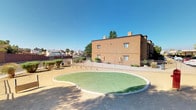 This single-level outdoor space presents a well-maintained community area characterized by its signature organic, kidney-shaped artificial turf installation, edged with a clean concrete border that contrasts attractively against the surrounding sandy desert-tone ground. The turf area serves as a central focal point, offering a lush green gathering space within an otherwise arid landscape setting.

The space is framed by warm earth-toned stucco buildings that reflect a Southwestern architectural style, complete with flat rooflines and minimal ornamentation typical of the region. Mature eucalyptus and other desert-adapted trees provide natural shade and visual depth throughout the area. A covered carport structure is visible to one side, offering sheltered vehicle parking for residents or visitors.

Security and definition of the space are provided by wrought iron fencing that encloses the perimeter, creating a safe and organized environment. The open-air design allows for abundant natural sunlight throughout the day, enhanced by the characteristically clear blue skies of the desert Southwest. The overall ambiance is clean, functional, and community-oriented, with a thoughtful balance between hardscape and greenery that makes it an inviting outdoor retreat suited to the surrounding neighborhood environment.
