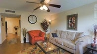 This single-floor space centers around a comfortable and thoughtfully arranged living room. The room is furnished with a spacious beige sofa accented by a decorative pillow, paired with a bold rust-red armchair that adds a pop of color and visual contrast. A glass-top coffee table with a metal frame anchors the seating arrangement, resting atop a patterned area rug that adds texture and warmth to the hardwood-style flooring beneath.

A ceiling fan with an integrated light fixture provides both air circulation and ambient lighting, complementing a tall floor lamp that adds a softer, secondary light source. Natural wall tones keep the space feeling open and neutral, serving as a backdrop for decorative accents including a large wall clock, an ornamental potted plant with dark foliage, and a framed artwork print. Dried botanicals in a tall vase contribute additional organic texture.

The overall ambiance is warm and lived-in, with a traditional aesthetic that balances comfort and style. The layout is practical and welcoming, making the living area a functional centerpiece of this single-floor home.
