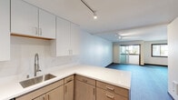 This well-appointed single-floor residence showcases a thoughtful, modern design that seamlessly blends functionality with contemporary style. The open-concept layout connects the kitchen and living area, creating a spacious and airy feel throughout.

The kitchen is a standout feature, outfitted with two-tone cabinetry — crisp white upper cabinets paired with warm wood-toned lower cabinets — and generous white quartz countertops that extend into a peninsula with additional storage. Stainless steel appliances, including a range with microwave, dishwasher, and refrigerator, complete the culinary space. A stainless steel sink with a modern pull-down faucet and a stainless steel backsplash behind the range add polished, practical touches throughout.

The living area is bathed in natural light, courtesy of a sliding glass door that opens to a private balcony, along with an additional window offering views of the surrounding building. Track lighting runs throughout the space, providing adjustable illumination across both the kitchen and living zones. Flooring transitions from wood-look planks in the main living area to plush blue-toned carpet near the balcony.

The bathroom is elegantly finished with travertine-style tile flooring, wood-framed doorways, and a glass-enclosed tub and shower combination featuring warm wood-toned wall tiling, lending a spa-like quality to the space.