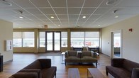 This single-floor communal lounge offers a welcoming and well-appointed environment designed for comfort and social interaction. The space features a suspended drop-tile ceiling with recessed lighting throughout, providing consistent, even illumination across the room. Warm hardwood flooring anchors the interior and adds a sense of warmth to the overall aesthetic.

Generous windows and roller shades line the walls, allowing abundant natural light to fill the space while offering flexibility in light control. A set of glass double doors opens directly onto an outdoor terrace, which is equipped with seating and furniture, making it ideal for outdoor gatherings. The terrace overlooks a scenic open landscape, extending the sense of space well beyond the interior.

The lounge is furnished with plush, earthy-toned upholstered sofas and armchairs arranged in conversational groupings across area rugs, creating inviting pockets for relaxation or informal meetings. Structural columns punctuate the space, subtly defining different seating zones without interrupting the open-plan layout.

Warm, neutral wall tones complement the natural wood flooring and soft furnishings, contributing to a calm and professional atmosphere. Overall, this space balances functionality with comfort, making it well-suited for use as a common lounge, event space, or gathering area.