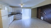 This single-floor space features an open-concept layout that seamlessly connects the kitchen and living area, creating a functional and cohesive environment. The kitchen is equipped with white cabinetry, dark countertops, and a white electric range with an overhead ventilation hood. The light-toned tile flooring extends throughout the space, providing a clean and unified look underfoot. A textured ceiling adds dimension overhead, while a ceiling fan with a built-in light fixture serves the living area, providing both airflow and illumination. A full-length sliding glass door dressed with vertical blinds floods the interior with natural light and provides access to the outdoors. The neutral beige wall tones complement the white cabinetry and bright tile, contributing to an airy and open ambiance. A solid wood entry door is visible along one wall, adding a warm, contrasting element to the otherwise light interior palette. The open layout maximizes the usable floor space, making the area feel spacious and versatile for everyday living.