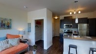 This single-floor unit features an open-concept design that seamlessly integrates the living, dining, and kitchen areas into one cohesive space. The kitchen is outfitted with stainless steel appliances, including a range and an over-the-range microwave, complemented by dark cabinetry that provides a modern contrast against the light-toned walls. A kitchen island with a countertop overhang offers additional prep space and doubles as a casual dining area, with bar-style seating. Dark hardwood flooring runs throughout, adding warmth and continuity to the space. The living area is comfortably sized and benefits from ambient lighting, with enough room to accommodate standard furnishings. A walk-in closet provides generous storage and includes built-in shelving and a hanging rod, offering practical organization options. Neutral wall tones keep the interior feeling bright and open, while recessed and track lighting fixtures provide functional illumination throughout. The overall ambiance is modern and functional, suited for everyday living.