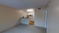 This single-floor residential space offers a practical and comfortable layout designed for everyday living. The living area is finished with neutral-toned wall-to-wall carpeting, creating a warm and understated ambiance throughout. A textured ceiling adds a classic touch to the interior.

The kitchen is accessible directly from the living area and is defined by a peninsula-style breakfast bar that serves as a natural divider between the two spaces. The kitchen features white cabinetry, white appliances including a refrigerator, electric range, and dishwasher, all set against hardwood-style flooring that contrasts with the carpeted living space. Under-cabinet lighting brightens the work area, and ample cabinetry provides generous storage.

A central HVAC vent is visible in the wall, ensuring climate control throughout the unit. The neutral color palette of soft beige and white tones across both spaces lends a clean, cohesive feel. The open sightlines between the kitchen and living area contribute to an airy, connected atmosphere, making the space feel welcoming and easy to navigate. Overall, the layout prioritizes functionality while maintaining a simple, inviting residential character.