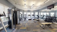 This single-floor fitness center is designed to accommodate a comprehensive range of workout activities within a spacious, open layout. The room is finished with cool blue-gray walls and crisp white trim, creating a clean and focused atmosphere conducive to exercise. Underfoot, a patchwork of multi-toned carpet tiles in shades of gray, blue, and yellow adds a contemporary visual element while providing comfort and durability throughout the space.

Natural light enters generously through multiple large, white-framed windows positioned along the walls, supplemented by suspended linear fluorescent fixtures and recessed lighting overhead. Ceiling fans are distributed throughout the room, contributing to air circulation and comfort during workouts.

The cardio zone is well-stocked with multiple treadmills and elliptical machines, each equipped with individual display screens. A dedicated strength training area includes a multi-station cable machine, free weights, a dumbbell rack, and weight benches. A wall-mounted flat-screen television is positioned for easy viewing from the workout floor.

Full-length mirrors are installed along one wall, visually expanding the space and allowing users to monitor their form. A yoga mat stored on the wall and additional floor space indicate provisions for stretching and mat-based exercises. The overall design balances functionality with a modern, well-maintained aesthetic.