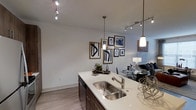 This single-floor space showcases a contemporary open-concept design that integrates the kitchen, dining, and living areas into one cohesive environment. The kitchen is anchored by a generous island topped with a light-toned countertop and fitted with a double stainless steel sink, complemented by rich dark cabinetry beneath. Overhead, a combination of track lighting and elegant pendant lights provides layered illumination, enhancing the clean, modern aesthetic throughout.

The light wood-style flooring runs continuously across the space, contributing to a sense of openness and warmth. Neutral-toned walls serve as a versatile backdrop, with a curated gallery wall of framed black-and-white artwork adding visual interest and personality to the dining area. The dining space accommodates a glass-top table, while the living area beyond features contemporary furnishings, completing the open layout.

The overall ambiance is sleek and understated, with thoughtful design choices—layered lighting, quality finishes, and cohesive décor—creating a space that feels both functional and stylishly refined.