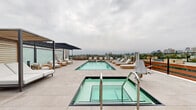 Located along Santa Monica Boulevard in Los Angeles, this rooftop amenity deck offers a sophisticated outdoor retreat with striking views of the city skyline and the Santa Monica Mountains. The centerpiece of the space is a generously sized lap pool finished with aqua-toned tile, surrounded by large-format, light-colored stone pavers that lend a clean, modern aesthetic throughout the deck. Adjacent to the pool sits a separate spa, complete with stainless steel handrails and an ADA-accessible pool lift, underscoring the space's commitment to inclusive design. Rows of teak-framed chaise lounges with crisp white cushions line both sides of the pool, providing ample space for sunbathing and relaxation. A bold, dark-framed pergola anchors one end of the deck, offering a shaded lounge area furnished with teak armchairs and side tables—ideal for more sheltered gatherings. A separate covered nook with additional daybed-style seating provides another intimate retreat beneath a slatted overhead structure. Glass panel railings along the perimeter ensure unobstructed panoramic views of the Los Angeles skyline and surrounding neighborhoods, making this rooftop deck a compelling and well-appointed outdoor destination.