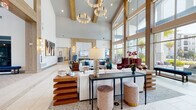 This expansive single-story communal lounge showcases a striking contemporary design defined by dramatically soaring ceilings accented with natural wood beam detailing. Exposed structural beams cross overhead, complemented by sculptural, geometric chandelier lighting that adds a refined focal point to the voluminous space. Large-format neutral tile flooring spans the entire area, reinforcing the clean, open aesthetic throughout.

Floor-to-ceiling window walls on multiple sides flood the interior with natural light while offering unobstructed views of manicured outdoor areas, including a pool courtyard framed by mature palm trees. Multiple seating vignettes are thoughtfully arranged throughout the space, featuring white upholstered sofas, bold blue and burgundy wingback accent chairs, layered wood-tiered coffee tables, and a sleek console table with velvet ottomans. A stone-clad fireplace serves as an elegant focal point in one section, paired with a mounted display screen and globe pendant sconces.

A separate café or refreshment bar area is visible toward the rear, adding functional versatility. The overall ambiance balances warmth and sophistication, combining natural wood tones, vibrant accent colors, and curated artwork to create an inviting, upscale communal environment.