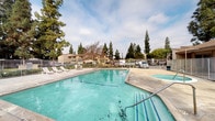 This outdoor community amenity space centers around a generously sized swimming pool with clear turquoise water, complemented by an adjacent circular spa. The pool deck is constructed of broad concrete, providing ample space for lounging and movement around the water. A set of white lounge chairs and a patio table are positioned poolside, offering a comfortable area for relaxation. The perimeter of the pool area is enclosed by a metal vertical-bar security fence, with a gated entry point providing controlled access to the space. A safety life ring is mounted along the interior fencing, ensuring emergency readiness. The surrounding landscape features tall, mature pine and deciduous trees that provide a natural backdrop and partial shade to the area. Residential buildings with a neutral earth-tone exterior are visible beyond the fence line, indicating this is a shared amenity within a multi-unit community. The open-air setting benefits from natural daylight and an expansive sky view. The concrete deck shows weathering consistent with an established outdoor space. Overall, the pool area offers a functional and accessible outdoor retreat for community residents.