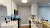 This single-floor unit presents a clean, contemporary aesthetic defined by neutral grey walls, wood-look plank flooring, and white trim throughout. The galley-style kitchen is efficiently laid out with white cabinetry, granite-look countertops, and stainless steel appliances including a gas range with overhead microwave and a dishwasher. A stainless steel sink anchors one side of the kitchen, while a pass-through peninsula connects the space to the main living area, enhancing the open feel. The living area is generously proportioned, featuring recessed lighting set into a textured ceiling and a sliding glass door that introduces natural light and provides access to an outdoor balcony area. The bedroom is a comfortable, well-defined space with a large window fitted with vertical blinds that allow for adjustable natural light. Mirrored sliding closet doors reflect light and add a sense of depth to the room. The wood-look flooring runs consistently throughout the unit, contributing to a cohesive and modern interior. Overall, the space offers a practical, well-organized layout with updated finishes suited for comfortable everyday living.
