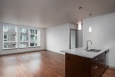 This well-appointed single-floor residence offers a thoughtfully designed open-concept layout that seamlessly connects the kitchen and living areas. The kitchen serves as a focal point, featuring rich dark-wood cabinetry, granite countertops, and a full suite of stainless steel appliances including a refrigerator, electric range, and built-in microwave. A central island with an integrated sink and pendant lighting above adds both functionality and visual appeal, while a tile backsplash and track lighting complete the polished look. Warm hardwood flooring extends throughout the main living areas, complementing the neutral gray accent walls and creating an inviting, cohesive atmosphere. The open living space offers a generous, flexible area bathed in natural light from large windows that frame views of the surrounding building complex. The bedroom continues the bright, airy aesthetic with plush carpeting, a large window, and a baseboard heating unit. The bathroom is finished with a dark-wood vanity, granite countertop, and a well-lit mirror, with the added convenience of a stacked washer and dryer tucked neatly within the same space. Clean lines, contemporary finishes, and an efficient floor plan make this residence a comfortable and functional modern living space.