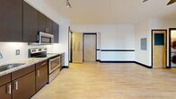 This one-bedroom apartment on the third floor of 50 W Fourth St in downtown Winston-Salem offers a well-designed open layout with light hardwood flooring and bold dark baseboards and door trim throughout. The modern kitchen features dark cabinetry reaching to the ceiling, granite countertops, stainless steel appliances including a range, microwave, dishwasher, and refrigerator, and track lighting overhead. The spacious bedroom is filled with natural light from multiple windows with urban views, and includes a ceiling fan. A closet and en-suite access are also present. The open living area flows seamlessly from the kitchen, creating an efficient, cohesive living space.