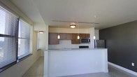 This single-floor space showcases a thoughtfully designed open-concept layout that seamlessly connects the kitchen and living areas. The kitchen is a standout feature, outfitted with rich, warm-toned wood cabinetry that provides ample storage, complemented by sleek stainless steel appliances including a built-in microwave and full-size refrigerator. A peninsula with a light granite countertop serves as both a functional workspace and a casual dining or entertaining surface, elegantly separating the kitchen from the adjoining living area. Overhead, a track lighting rail runs the length of the kitchen, paired with stylish elongated pendant lights that cast a warm, focused glow over the workspace. The living area features neutral-toned carpeting that adds softness underfoot, while windows fitted with blinds allow for adjustable natural light throughout the day. The walls are finished in a clean, neutral palette that enhances the sense of openness and brightness throughout the space. Flush-mount ceiling fixtures provide additional ambient lighting. The overall ambiance is clean, modern, and functional, making it well-suited for everyday living and casual entertaining alike.