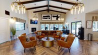 Located at 3980 W Linda Vista Blvd in Tucson, Arizona, this single-story community clubhouse offers a thoughtfully designed gathering space that blends modern Southwest aesthetics with functional amenities. The main lounge is anchored by soaring vaulted ceilings with exposed dark wood beams, herringbone-patterned wood-look flooring, and a striking stacked-stone fireplace topped by a wall-mounted TV. Cognac leather armchairs surround a gold drum coffee table, creating a welcoming seating arrangement bathed in natural light from large windows that overlook the outdoor pool area. Pendant chandeliers with Edison-style bulbs add a warm, industrial-chic touch throughout. A long, white quartz co-working counter with mid-century modern wood chairs provides ample workspace, illuminated by views of the pool through wide windows and accessed via French doors to the outdoor patio. Bold abstract artwork adorns the walls, complementing the sophisticated decor. Adjacent to the lounge, a dedicated coffee bar features a geometric tile accent wall, built-in cabinetry, a beverage refrigerator, and bistro-style seating at gold-topped tables. The fitness center is well-appointed with treadmills, ellipticals, strength training equipment, floor-to-ceiling mirrors, rubber flooring, and vibrant desert landscape artwork, completing this comprehensive community facility.