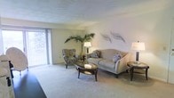 This single-floor space offers a comfortable and elegantly designed living environment. The living room serves as the centerpiece of the home, featuring plush wall-to-wall carpeting in a neutral tone that complements the soft, light-colored walls. Large vertical-blind-covered windows allow natural light to filter into the space, creating a warm and inviting atmosphere.

The living area is thoughtfully arranged with a classic sofa adorned with decorative pillows, a coordinating patterned armchair, and a rich, dark-wood oval glass-top coffee table that anchors the seating arrangement. A matching side table adds to the cohesive aesthetic. Decorative metal leaf wall art adds an artistic touch to the walls, while a tall indoor palm plant introduces a natural, organic element to the room.

The ceiling features a subtle textured finish, adding a layer of visual interest overhead. The overall design palette is neutral and understated, with layered textures and carefully selected furnishings that create a sense of understated sophistication. The space feels open and airy, with a layout that encourages comfortable living and relaxed gatherings. The cohesive blend of soft furnishings, natural elements, and tasteful décor gives the space a polished yet livable character.
