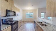This single-floor residence presents a thoughtfully designed open-concept layout that connects the kitchen, dining, and living areas into one cohesive space. The kitchen is well-appointed with light wood cabinetry, speckled granite countertops, black appliances including a range, microwave, and refrigerator, and a double basin sink. A peninsula with a granite overhang provides casual seating and visually separates the kitchen from the adjoining living area. Natural light enters through multiple windows, complemented by a chandelier over the dining space and a flush-mount fixture in the kitchen corridor. Light-toned luxury vinyl plank flooring runs throughout the main living areas, lending a clean and cohesive aesthetic. The bedrooms are comfortably sized, with large windows offering outdoor views; one room features a ceiling fan fixture. The unit includes two bathrooms: one with a walk-in shower stall and granite-topped vanity, and another with a tub-shower combination. A dedicated laundry area with washer and dryer hookups is visible through the kitchen corridor, adding functional convenience. The overall palette of warm neutrals and natural wood tones creates a bright, welcoming atmosphere.