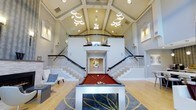 This single-story clubhouse makes a striking impression with its dramatic vaulted ceiling, which rises high above the main gathering area and is adorned with a series of contemporary circular pendant light fixtures that cast a warm, welcoming glow throughout the space. The open-plan layout centers around a bold red pool table set atop a decorative area rug with an artistic botanical pattern, establishing the room as a premier social and recreational destination.

Twin symmetrical staircases flank the rear of the space, each featuring sleek metal railings with dark wood handrails, and they curve gracefully up to an elevated mezzanine-style overlook that adds architectural depth and visual interest to the interior. The space beneath this upper landing connects to additional rooms beyond, hinting at further amenities within the facility.

Light hardwood flooring runs throughout, complementing the neutral gray and cream wall tones and contributing to the overall brightness of the space. Decorative wall art, built-in architectural molding details, and comfortable accent seating along the perimeter complete the polished, upscale atmosphere of this well-appointed clubhouse.