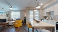 This single-floor residence offers a thoughtfully designed open-concept layout that seamlessly connects the kitchen, dining, and living areas. The kitchen is outfitted with crisp white cabinetry, a subway tile backsplash, stainless steel appliances including a refrigerator, range, and over-the-range microwave, and a white quartz-topped island illuminated by dual industrial-style pendant lights. Rich hardwood-style flooring runs throughout the main living areas, adding warmth and continuity to the space. The dining area flows naturally into the living room, which is anchored by a sleek electric fireplace framed by a clean white mantle. Large windows dressed with sheer curtains and horizontal blinds allow abundant natural light to fill the space, while a ceiling fan adds comfort overhead. A set of glass French doors provides access to the outdoors. The bedroom is a tranquil retreat featuring plush carpeting, a large window with sheer window treatments, and ample natural light. The en-suite bathroom is finished with a white vanity, wood-style flooring, and a walk-in shower adorned with large-format marble-look tile and a rainfall showerhead, delivering a clean and contemporary aesthetic throughout.
