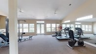 This single-story fitness room offers a well-rounded exercise environment designed to support a variety of workout routines. The space is generously sized, providing ample room for both cardio and strength training activities. Large, multi-pane windows line the walls, flooding the interior with abundant natural light and offering views of the surrounding greenery. A set of French doors with transom windows serves as the primary entrance, enhancing the bright and open atmosphere. Ceiling fans are installed overhead, promoting air circulation throughout workouts. The flooring is covered in low-pile carpet, offering a comfortable and practical surface underfoot. Equipment includes treadmills and an elliptical machine positioned along one wall, alongside a weight bench and cable machine for strength training. A large wall-mounted mirror on one side of the room visually expands the space and allows users to monitor their form during exercise. The walls are finished in a neutral beige tone, contributing to a clean and functional aesthetic. Overall, the fitness room presents a welcoming and practical environment well-suited for daily exercise.