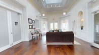 This single-floor space makes an immediate impression with its dramatic ceiling height, anchored by a recessed skylight fitted with track lighting that bathes the room in a combination of natural and artificial light. Crown molding runs along the upper walls, reinforcing the refined architectural character of the space. Large transom windows positioned high on the walls further enhance the sense of openness and brightness throughout.

The main living area centers around a substantial dark chocolate sectional sofa resting on a neutral area rug, complemented by a pair of elegant side chairs and a glass-topped accent table in a dedicated seating nook. A decorative glass block feature wall adds a distinctive design element, while a small cluster of framed artwork and fresh floral accents contribute warmth to the otherwise crisp, neutral palette.

Wide-plank hardwood flooring in a rich, warm tone spans the entire space, adding texture and depth. French doors along the far wall open to an exterior view, allowing abundant natural light and a visual connection to the outdoors. The overall ambiance is polished and welcoming, combining classic architectural details with comfortable furnishings.