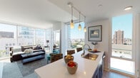 This contemporary urban residence is thoughtfully designed to maximize natural light and city views, with floor-to-ceiling windows and glass doors that frame striking skyline panoramas from nearly every room. The open-concept layout seamlessly connects the kitchen, dining, and living areas, creating a bright and cohesive living space finished with light hardwood-style flooring throughout.

The kitchen is a standout feature, equipped with dark cabinetry, stainless steel appliances including a refrigerator, microwave, and range, a subway tile backsplash, and a large central island with a white quartz countertop and undermount sink. Pendant lighting over the island adds warmth and definition to the space. Bar seating at the island makes it ideal for casual dining.

The living area is anchored by expansive corner windows that flood the room with natural light and offer unobstructed views of the surrounding cityscape, including notable architectural landmarks. Access to an outdoor balcony further extends the living space.

The residence includes two well-appointed bedrooms, each with closet storage and large windows overlooking the city. Both bathrooms feature dark vanity cabinetry, light tile flooring, and illuminated mirrors, maintaining the clean, modern aesthetic found throughout the home. The overall ambiance is sophisticated, contemporary, and urban.