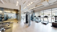 Located at 645 E Ocean Blvd in Long Beach, California, this fitness center offers a thoughtfully designed, single-floor space that balances functionality with a clean, modern aesthetic. The open layout is defined by light hardwood-style flooring, exposed ductwork on high white ceilings, and an abundance of natural light streaming through floor-to-ceiling glass windows that frame views of the surrounding streetscape.

Pendant globe lights suspended from the ceiling provide warm, even illumination throughout the space, complementing the industrial-modern architectural character. The main workout area is well-equipped with multiple treadmills, elliptical machines, stationary and spin bikes, a stair climber, and a multi-functional cable machine, offering a comprehensive range of cardio and strength training options.

A dedicated free weights zone features a full dumbbell rack, weight benches, and exercise mats positioned in front of a large wall mirror, ideal for resistance training and stretching. A glass-enclosed area provides additional workout space, visible from the main floor.

Thoughtful amenities include built-in shelving with a mounted television, a water station, and locker access, contributing to a well-rounded fitness experience. The overall ambiance is bright, airy, and professionally appointed, making this fitness center a premium facility for residents and users of the Ocean Blvd address.