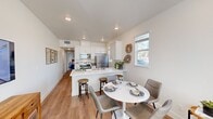 This single-story home makes excellent use of its footprint, accommodating four bedrooms, one bathroom, and a combined living, dining, and kitchen area. The open-concept main living space is bright and airy, featuring warm wood-tone flooring, recessed lighting, and a neutral white color palette that creates a clean, modern aesthetic. The kitchen is well-appointed with white shaker cabinetry, crisp white quartz countertops, a peninsula with bar seating, and a full suite of stainless steel appliances including a range, over-the-range microwave, and refrigerator. A window above the sink allows natural light to fill the space. The dining area adjoins the kitchen and flows seamlessly into the living room, which features a large window with blinds that brings in additional natural light. The primary bedroom offers sliding glass door access to a private outdoor patio area, providing a direct indoor-outdoor connection. Additional bedrooms are compact and functional, each with windows that allow natural light and views of the surrounding outdoor space. The shared bathroom includes a glass-enclosed shower stall with white cabinetry for storage. Throughout the home, consistent light wood flooring and a bright, neutral palette unify the design.