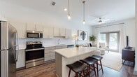 This single-story residence is designed with a modern, open-concept layout that seamlessly connects the kitchen, dining, and living areas into one cohesive, light-filled space. The kitchen is a standout feature, outfitted with two-tone cabinetry—upper white cabinets paired with textured gray lower cabinets—white quartz countertops, a subway tile backsplash, and a full suite of stainless steel appliances including a refrigerator, range, microwave, and dishwasher. A central island with bar seating serves as both a functional workspace and a casual dining spot, illuminated by stylish pendant lighting suspended from elevated ceilings. Rich wood-look flooring flows throughout the main living areas, grounding the bright, neutral palette. The living room is bathed in natural light through large windows and a glass door, complemented by a ceiling fan and access to outdoor views. The bedroom is a calm retreat, featuring a generous window with sheer drapery, wood-look flooring, and soft neutral tones. The bathroom continues the contemporary theme with wood-look flooring, a tub-shower combination, and clean white finishes throughout.