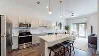 This single-story residence is designed with a modern, open-concept layout that seamlessly connects the kitchen, dining, and living areas into one cohesive, light-filled space. The kitchen is a standout feature, outfitted with two-tone cabinetry—upper white cabinets paired with textured gray lower cabinets—white quartz countertops, a subway tile backsplash, and a full suite of stainless steel appliances including a refrigerator, range, microwave, and dishwasher. A central island with bar seating serves as both a functional workspace and a casual dining spot, illuminated by stylish pendant lighting suspended from elevated ceilings. Rich wood-look flooring flows throughout the main living areas, grounding the bright, neutral palette. The living room is bathed in natural light through large windows and a glass door, complemented by a ceiling fan and access to outdoor views. The bedroom is a calm retreat, featuring a generous window with sheer drapery, wood-look flooring, and soft neutral tones. The bathroom continues the contemporary theme with wood-look flooring, a tub-shower combination, and clean white finishes throughout.
