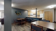 This single-floor communal space is designed to serve as a shared gathering and activity area. The layout is open and functional, seamlessly connecting a fully equipped kitchen, multiple dining areas, and a comfortable lounge zone. The kitchen features light wood cabinetry, a full-size refrigerator, electric range with overhead microwave, a double sink, and a wraparound peninsula counter with bar-height seating — ideal for communal meal preparation and casual dining. The flooring transitions between tile in the kitchen and wood-look vinyl plank throughout the common areas, adding warmth and cohesion. The dining area accommodates multiple rectangular tables paired with wood-framed chairs upholstered in dark faux leather, with decorative wall art adding visual interest. A separate lounge area is furnished with navy blue sofas, a rustic wooden coffee table, and accent chairs arranged around a flat-screen television, creating a relaxed setting for socializing. Large windows along one wall allow natural light to fill the lounge space. Textured ceilings and a muted color palette of slate blue and warm gray tones contribute to a welcoming, residential ambiance throughout.