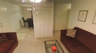 This single-floor space offers an open, functional layout that connects the living and dining areas seamlessly. The living area is anchored by a plush dark sofa accented with decorative throw pillows, positioned alongside a glass-top coffee table with a wrought-iron frame, adding a classic touch to the space. Soft, wall-to-wall carpeting runs throughout, contributing to a warm and comfortable ambiance.

A prominent set of louvered bi-fold closet doors serves as a central architectural feature, providing generous storage capacity while adding visual texture to the room. The ceiling is fitted with a light fixture, ensuring adequate illumination across the living space.

Adjacent to the living area, the dining space accommodates a dark-toned dining set with multiple chairs, illuminated by a chandelier overhead that lends a touch of elegance to everyday meals. The walls are finished in a neutral green-grey tone, creating a calm and cohesive atmosphere throughout. The overall layout is practical and well-proportioned, making efficient use of the available space across both living and dining zones.