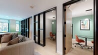 Located at 5 Port Imperial Blvd in Weehawken, NJ, this thoughtfully designed amenity space occupies a single floor and offers a refined blend of collaborative and private environments. The open lounge area serves as the heart of the space, featuring light hardwood flooring, a large wall-mounted display, and a curated mix of seating including a plush sofa, leather accent chairs, and a fluted wood coffee table, all anchored by a striking area rug. Sage green accent walls and abundant natural light from large windows create a warm, inviting atmosphere. The adjacent dining area provides a comfortable setting for casual gatherings, with contemporary furniture and continued access to natural light. A dedicated wet bar station with warm wood cabinetry, dark countertops, and a bold geometric patterned tile backsplash adds a functional yet stylish touch near the entry. Private meeting rooms, enclosed by sleek black-framed glass partition walls with wood-paneled doors, offer focused work settings. Each meeting room is furnished with a round or rectangular glass table, terracotta-toned upholstered chairs, pendant lighting, and sage green accent walls adorned with framed artwork, creating a cohesive and professional aesthetic throughout.
