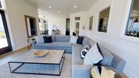 This single-floor space showcases a thoughtfully designed open-concept layout that seamlessly connects the living and dining areas, creating a sense of spaciousness and ease of flow throughout. The living area is anchored by a generous sectional sofa in soft blue-gray upholstery, accented with navy and geometric-patterned throw pillows that lend a modern coastal character to the room. A substantial square coffee table with a metal frame sits atop a layered area rug, grounding the seating arrangement with a relaxed yet refined aesthetic. Recessed lighting is installed throughout the ceiling, providing even, adjustable illumination that enhances the crisp white walls and bright atmosphere of the space. An open passage leads to the dining area, where pendant lighting hangs above what appears to be a dark-toned dining set, complementing the overall palette. Built-in dark cabinetry adds a striking contrast against the white walls, offering both visual interest and functional storage. The space benefits from natural light filtering through multiple windows and glass doors, contributing to the overall airy and welcoming ambiance. The clean lines, neutral tones, and carefully selected furnishings combine to create an environment that feels both contemporary and comfortable.