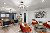 Located at 6565 McCallum Blvd in Dallas, Texas, this thoughtfully designed single-floor space serves as a welcoming office or leasing lobby, offering a cohesive blend of modern and eclectic interior design. The open layout is anchored by light gray wood-look flooring throughout, providing a clean, contemporary foundation that ties the entire space together.

The space is divided into two distinct yet connected zones. A comfortable lounge area greets visitors with a set of plush burnt-orange velvet armchairs arranged around a round metal coffee table, set atop a patterned area rug. Black-framed windows along the exterior walls flood the space with natural light and offer views to the outside. Bold abstract wall art adds a vibrant artistic touch.

Opposite the lounge, a reception and workspace area features a striking wood-and-textured-panel desk with leather seating, set against a deep navy accent wall bearing a welcoming "HELLO" sign. A round decorative mirror and a wooden console table with curated décor complete this zone's refined aesthetic.

A private office at the rear showcases a substantial wooden executive desk illuminated by a sculptural metal ring pendant light — one of several statement fixtures suspended throughout the space. Recessed lighting supplements the pendants, ensuring the entire area is well-lit. Teal-painted doors and dark-framed glass partitions add purposeful contrast against the crisp white walls, creating a polished, professional ambiance.