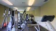This fitness center is a dedicated exercise space designed to support a well-rounded workout routine. The room is furnished with a selection of cardio equipment, including multiple treadmills, providing ample options for aerobic training. A multi-function strength training machine anchors one side of the space, offering users a range of resistance exercises in a compact footprint. Floor-to-ceiling mirrors line the walls, creating a sense of expanded space while allowing users to monitor their form during workouts. The walls are painted in a soft, neutral tone that contributes to a clean and focused atmosphere. Overhead fluorescent lighting ensures the space is well-lit throughout. A vending machine is visible in the background, adding a convenient amenity for post-workout refreshments. The flooring appears to be a durable, low-pile carpet suited for fitness use. A window allows natural light to filter into the room, enhancing the overall brightness of the environment. An instructional exercise chart is posted on the wall, offering guidance for users. The space is thoughtfully laid out to maximize usability while maintaining clear pathways between equipment.