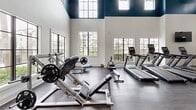 This expansive 1,740 square foot fitness center, situated at 11481 Northwest 41st Street in Doral, Florida, offers a comprehensive workout experience. The facility boasts high ceilings, creating an open and airy atmosphere enhanced by large windows that flood the space with natural light. The modern interior design features a harmonious blend of white walls and teal accents, complemented by durable wood-look flooring.

The gym is equipped with a wide array of exercise machines, including cardio equipment such as treadmills and ellipticals, as well as strength training apparatus like weight machines and free weights. A multi-functional cable machine serves as a centerpiece, allowing for varied workout routines. The layout is thoughtfully organized, with distinct areas for different types of exercises.

Amenities include ceiling fans for air circulation, recessed lighting, and strategically placed mirrors to assist with form during workouts. The space also features a separate room with additional equipment, likely for group classes or specialized training. A glass door entrance provides easy access and visibility. This well-appointed fitness center caters to diverse fitness needs in a stylish, functional environment.