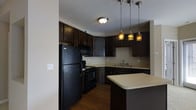 Located at 747 Worthington Woods Blvd in Worthington, Ohio, this single-floor residence offers a well-designed living environment with thoughtfully chosen finishes throughout. The kitchen serves as a standout feature, showcasing rich, dark espresso-toned cabinetry that extends from floor to ceiling, providing generous storage space. Black appliances, including a refrigerator, range, and built-in microwave, complement the bold cabinetry and create a cohesive, modern aesthetic. Light-toned countertops offer a striking contrast against the dark lower and upper cabinets, adding visual balance to the space. A peninsula with matching countertop extends into the room, offering additional workspace and casual dining or entertaining opportunities. Three decorative pendant lights hang above, casting warm, ambient light that enhances the kitchen's inviting atmosphere. The flooring features rich, dark hardwood-style planks that carry the sophisticated color palette throughout. The neutral wall tones keep the space feeling open and allow the cabinetry and fixtures to take center stage. A sink with modern fixtures is positioned along the far counter wall, completing this functional and stylish kitchen layout.
