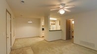 This single-floor residence features an open-concept layout that connects the living area and kitchen, creating a cohesive and functional space. The living area is finished with neutral carpet and is anchored by a ceiling fan with a built-in light fixture, providing both airflow and ambient lighting. A pendant light near the kitchen adds a decorative touch and helps define the transition between spaces.

The kitchen is positioned centrally and features a peninsula with a dark countertop, providing a natural dividing point between the cooking and living zones. White cabinetry lines the walls, complemented by light wood upper cabinets near the refrigerator. A full-size white refrigerator and additional appliances, including a microwave, are visible, indicating a well-equipped cooking space. Under-cabinet fluorescent lighting illuminates the work surfaces.

A hallway accessible from the main living area leads to what appears to be a bathroom and additional rooms, offering a degree of privacy from the common areas. The overall color palette is neutral throughout, with beige walls and light flooring, giving the space a clean and versatile foundation suitable for a variety of interior styles.