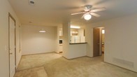 This single-floor residence features an open-concept layout that connects the living area and kitchen, creating a cohesive and functional space. The living area is finished with neutral carpet and is anchored by a ceiling fan with a built-in light fixture, providing both airflow and ambient lighting. A pendant light near the kitchen adds a decorative touch and helps define the transition between spaces.

The kitchen is positioned centrally and features a peninsula with a dark countertop, providing a natural dividing point between the cooking and living zones. White cabinetry lines the walls, complemented by light wood upper cabinets near the refrigerator. A full-size white refrigerator and additional appliances, including a microwave, are visible, indicating a well-equipped cooking space. Under-cabinet fluorescent lighting illuminates the work surfaces.

A hallway accessible from the main living area leads to what appears to be a bathroom and additional rooms, offering a degree of privacy from the common areas. The overall color palette is neutral throughout, with beige walls and light flooring, giving the space a clean and versatile foundation suitable for a variety of interior styles.
